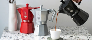 A lineup of 3 Bialetti Moka pots on a benchtop. Each Moka pots is a different colour (Red, Silver, and Black). Coffee has been brewed in the Black Moka Pot, and someone is pouring the coffee into a white espresso cup.